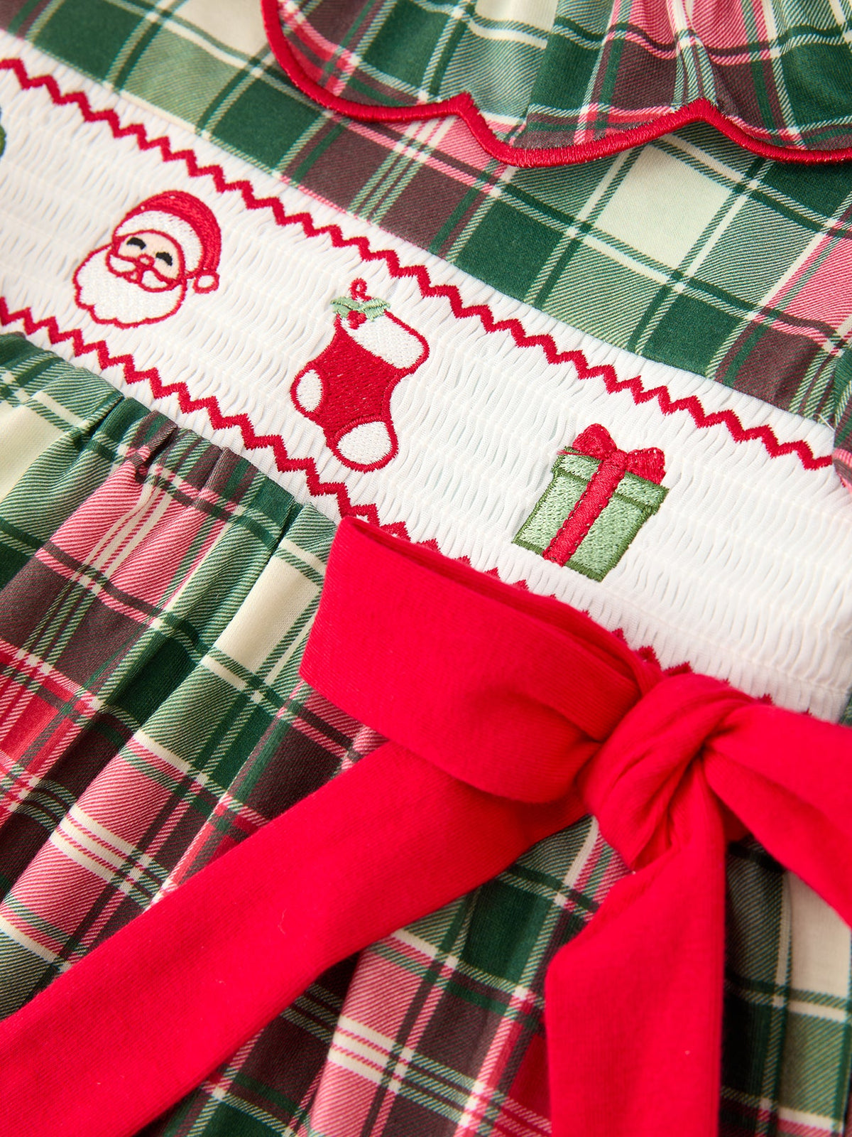 Girls Christmas plaid dress with smocked chest featuring embroidered Santa, stocking, and gift box, accented by a bold red bow.