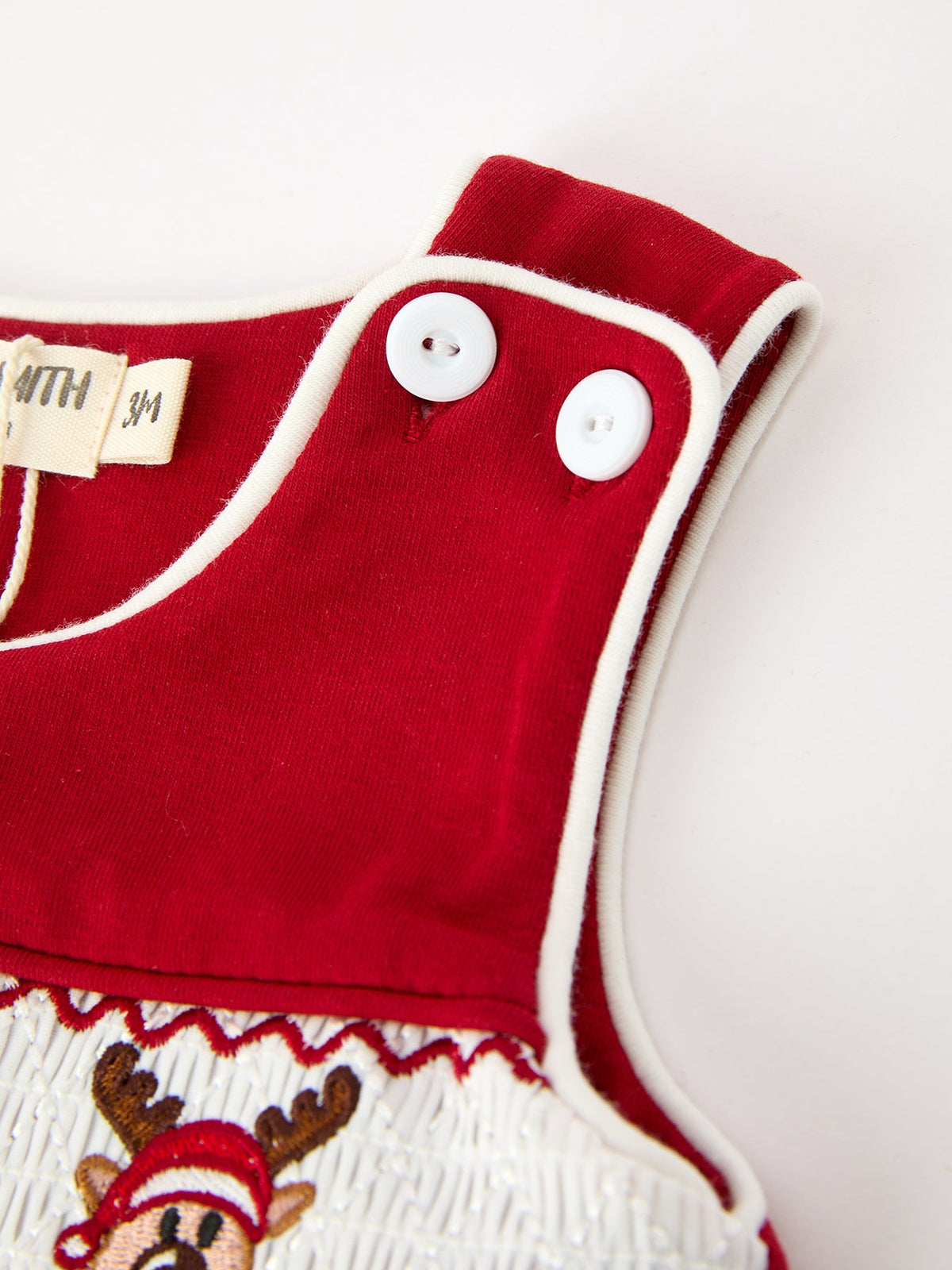 Red baby romper shoulder with white buttons, white contrast piping, and embroidered reindeer on smocked chest panel.