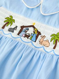 Blue baby romper features embroidered nativity scene on smocked chest with palm trees, donkey, and camel.