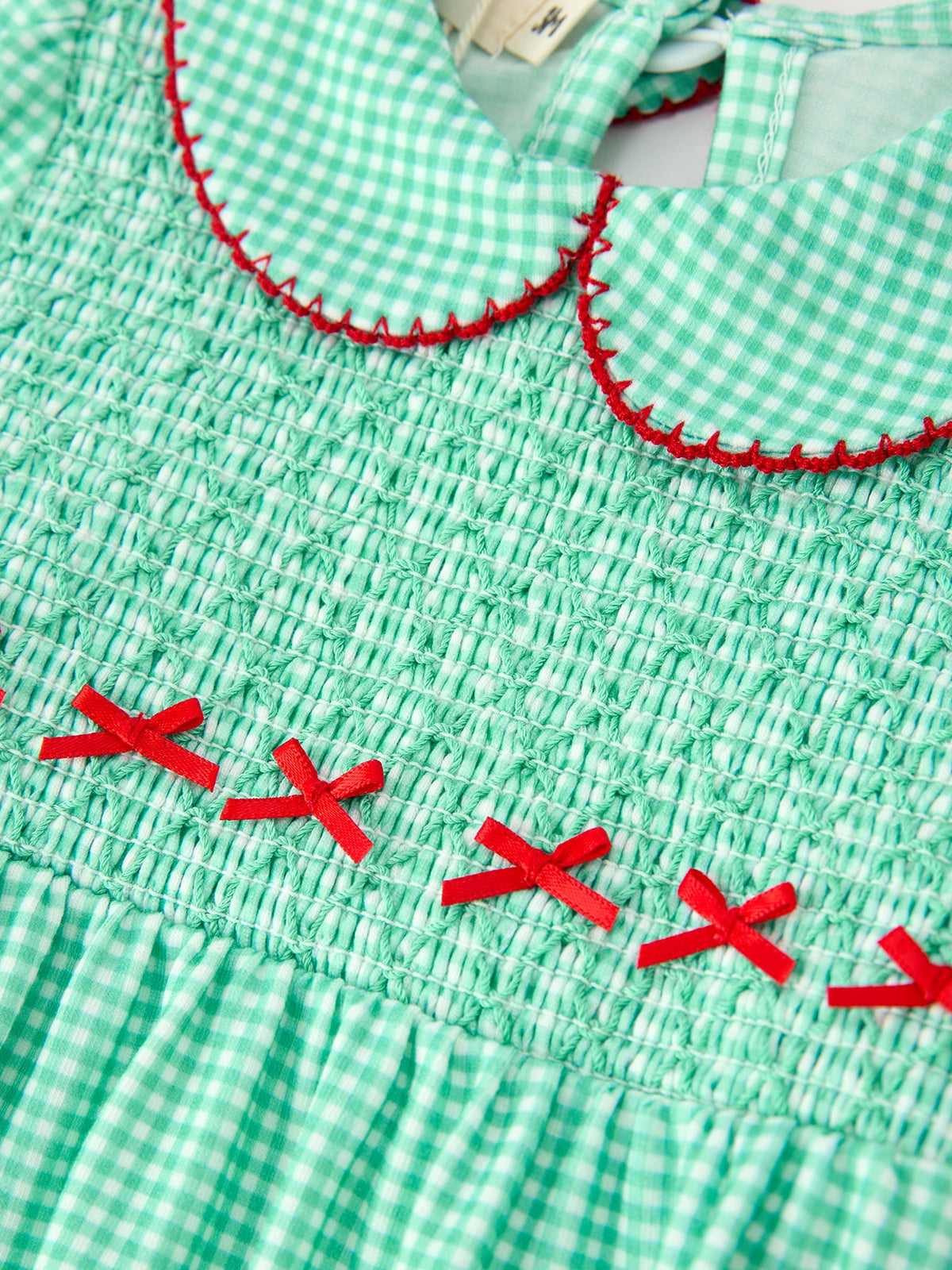 Green plaid smocked baby romper bodice with red stitched Peter Pan collar and small red ribbon bows for festive holiday wear.
