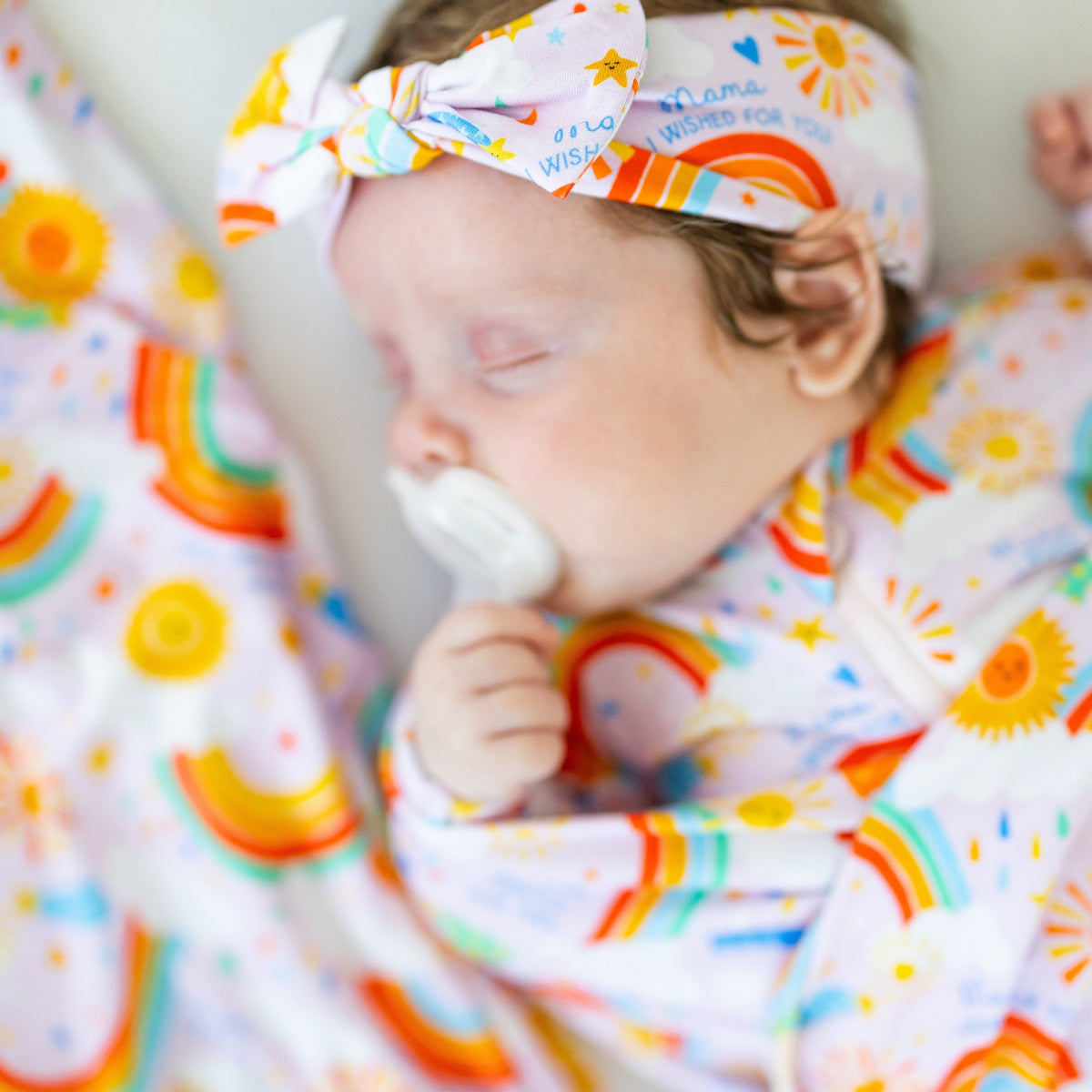 Baby wearing The Forever Swaddle Blanket - Here Comes the Sunshine (Pink) with a matching headband, enjoying a cozy moment while sucking on a pacifier, epitomizing comfort and cuteness.