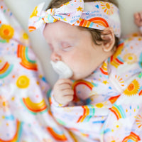 Baby wearing The Forever Swaddle Blanket - Here Comes the Sunshine (Pink) with a matching headband, enjoying a cozy moment while sucking on a pacifier, epitomizing comfort and cuteness.
