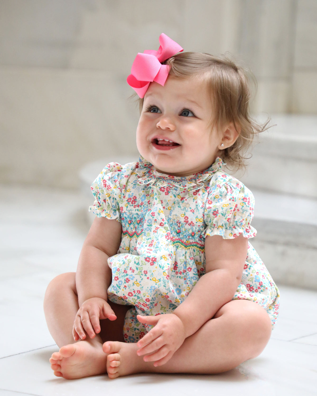 Baby sitting in floral bubble romper wearing large bright pink bow headband