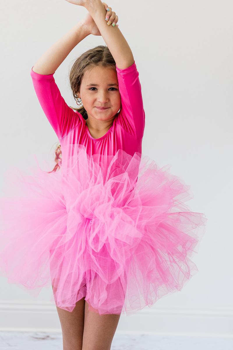 A young girl wearing a Hot Pink 3/4 Sleeve Fluffy Tutu Leotard for Girls, joyfully dancing with her arms raised. The vibrant tutu leotard for girls features multiple layers of soft tulle, creating a delightful fluffy appearance.