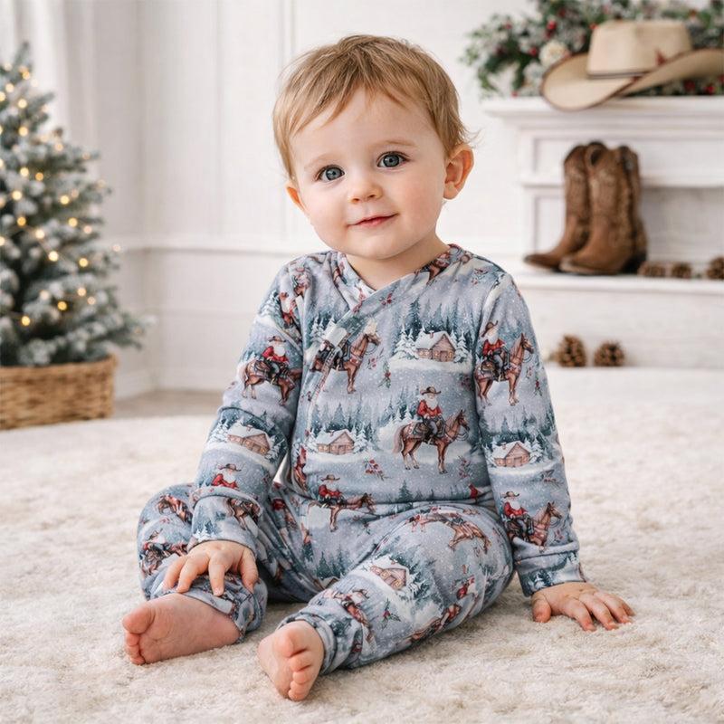 Baby in grey Santa print bamboo romper sitting on white rug near Christmas tree
