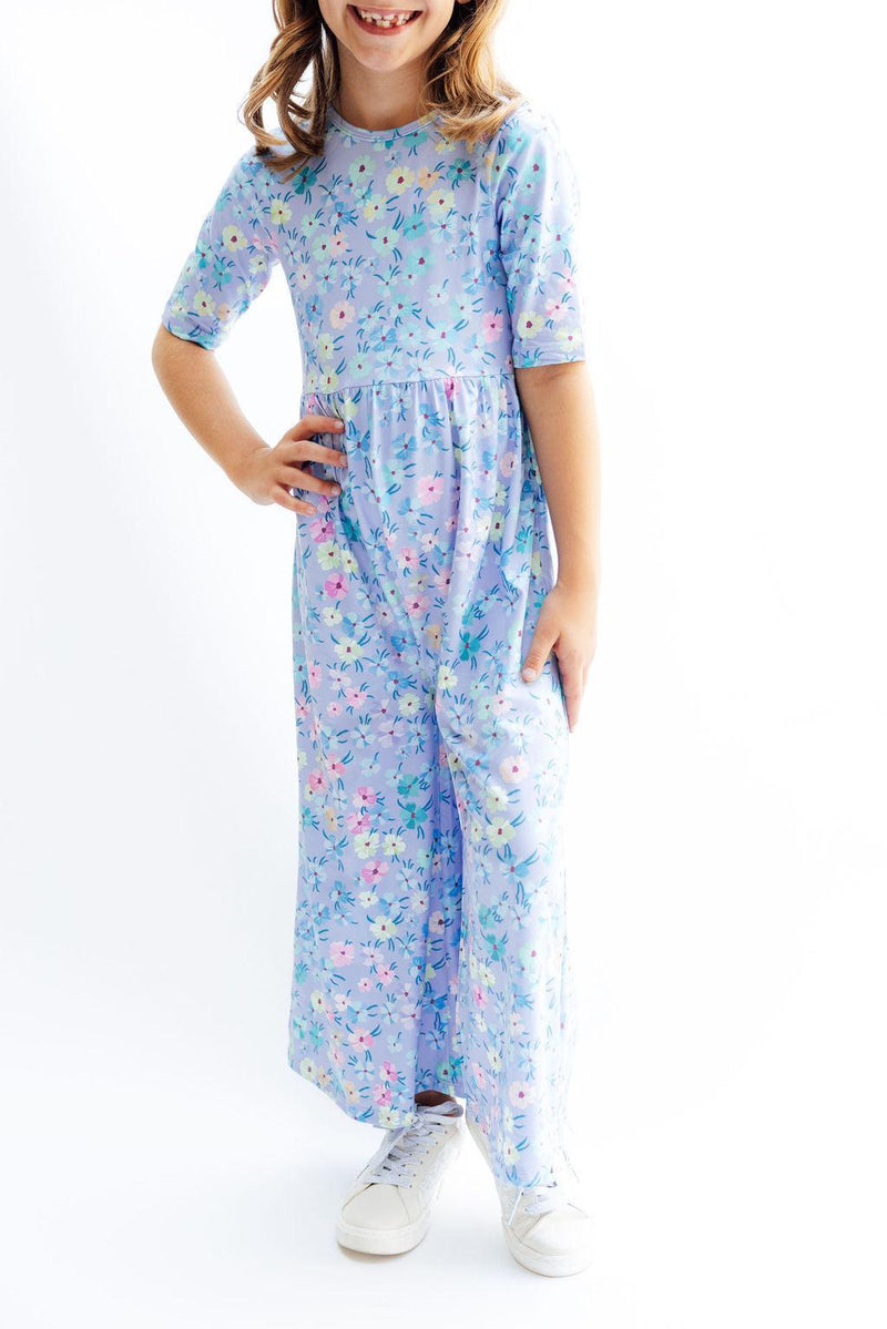 A young girl wearing the Petal Party Wide Leg Jumpsuit by Mila & Rose, smiling and posing with her hands on her hips. The jumpsuit features a light blue floral design, perfect for playdates.