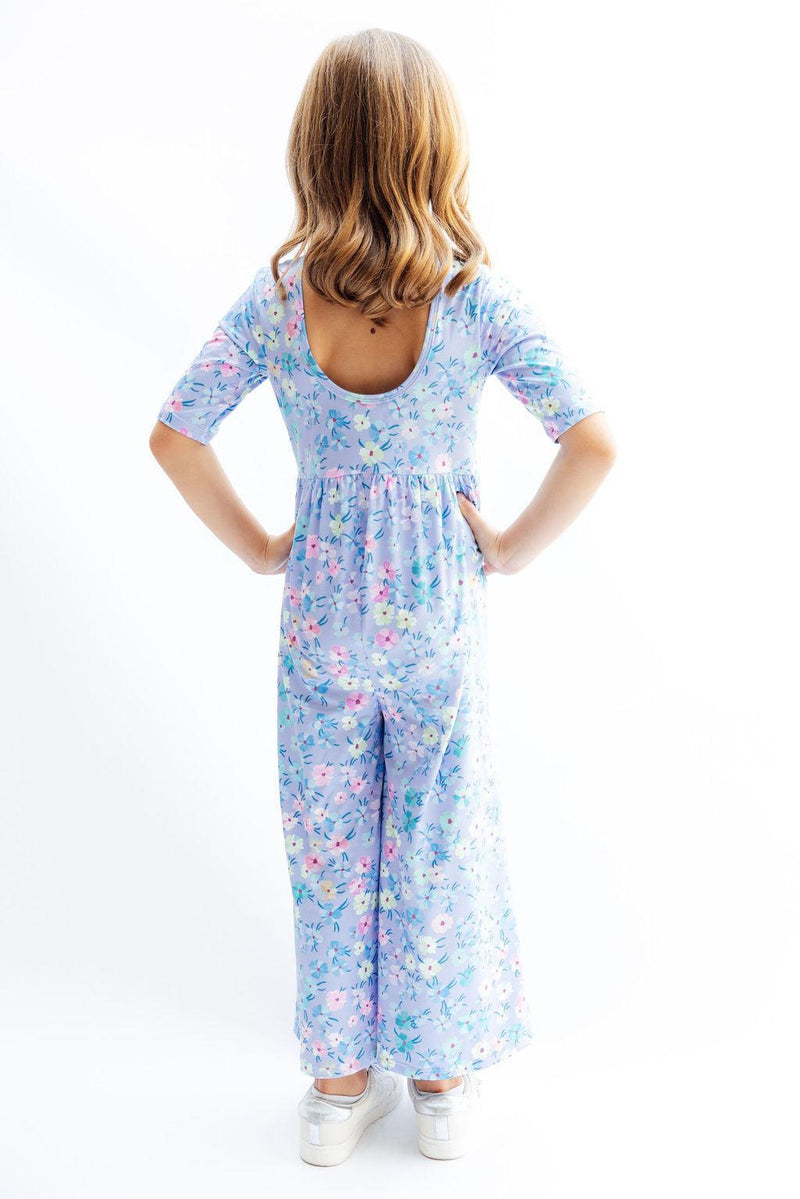 A young girl poses in the back view of the Petal Party Wide Leg Jumpsuit by Mila & Rose, showing off the unique scoop back design and colorful floral print.