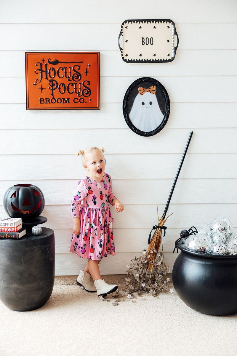 A joyful little girl wearing the Purr-fectly Spooky 3/4 Sleeve Pocket Twirl Dress, surrounded by Halloween decor, including signs and a broom, showcasing a fun and playful atmosphere.
