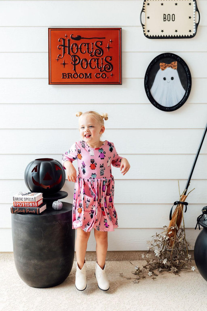 Happy girl in the Purr-fectly Spooky 3/4 Sleeve Pocket Twirl Dress, standing near Halloween decor, exuding joy and showcasing her stylish outfit.