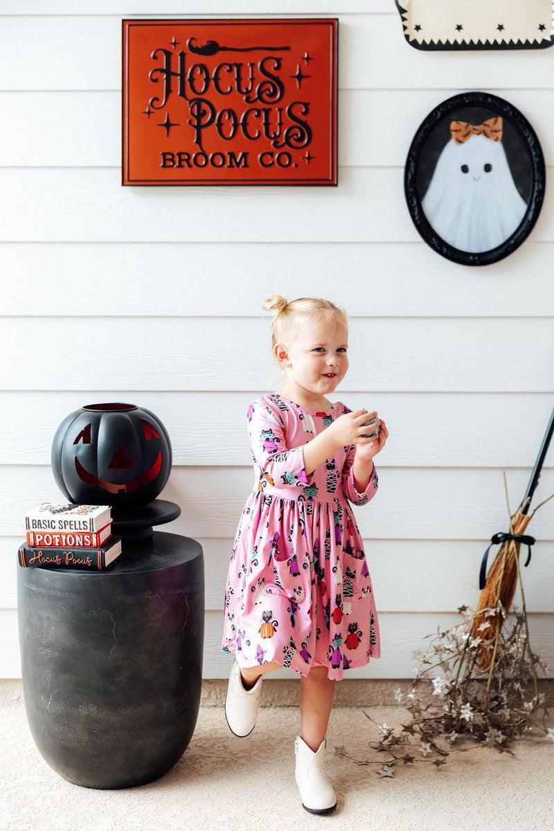 A young girl in the Purr-fectly Spooky 3/4 Sleeve Pocket Twirl Dress, playfully posing beside Halloween decorations like a jack-o'-lantern, highlighting the dress's fun personality.