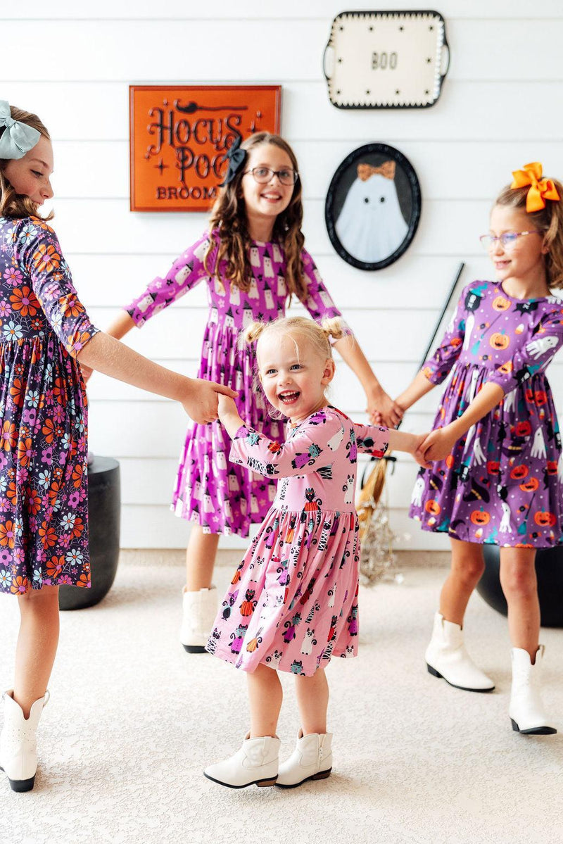 Children joyfully twirling in the Purr-fectly Spooky 3/4 Sleeve Pocket Twirl Dress, a fun Halloween attire by Mila & Rose. They are holding hands and smiling, showcasing the dress's playful design.