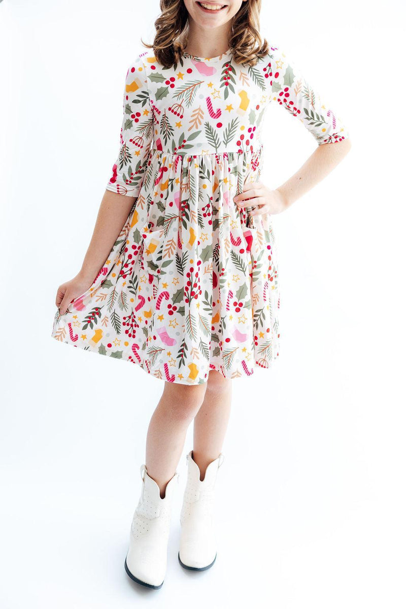 A full-length view of a girl wearing the 'Holly Jolly Christmas' 3/4 Sleeve Pocket Christmas Twirl Dress for Girls, paired with stylish white boots.