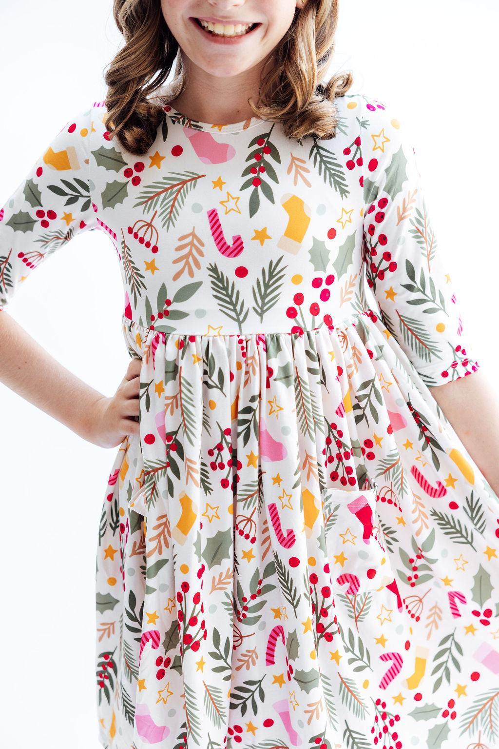 A young girl smiling while modeling the 'Holly Jolly Christmas' 3/4 Sleeve Pocket Christmas Twirl Dress for Girls, featuring a festive print and playful twirl design.