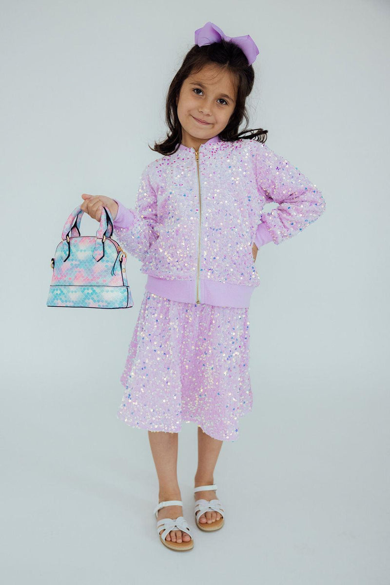 A young girl showcasing the 'Lilac Velvet Sequin Jacket' from Mila & Rose®, standing confidently with a sparkle in her eyes, complemented by a matching outfit and handbag, perfect for special occasions.