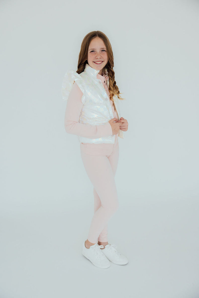 A girl dressed in Zen Pink Active Legging by Mila & Rose, holding a shiny iridescent vest, smiling confidently in a bright, simple setting.