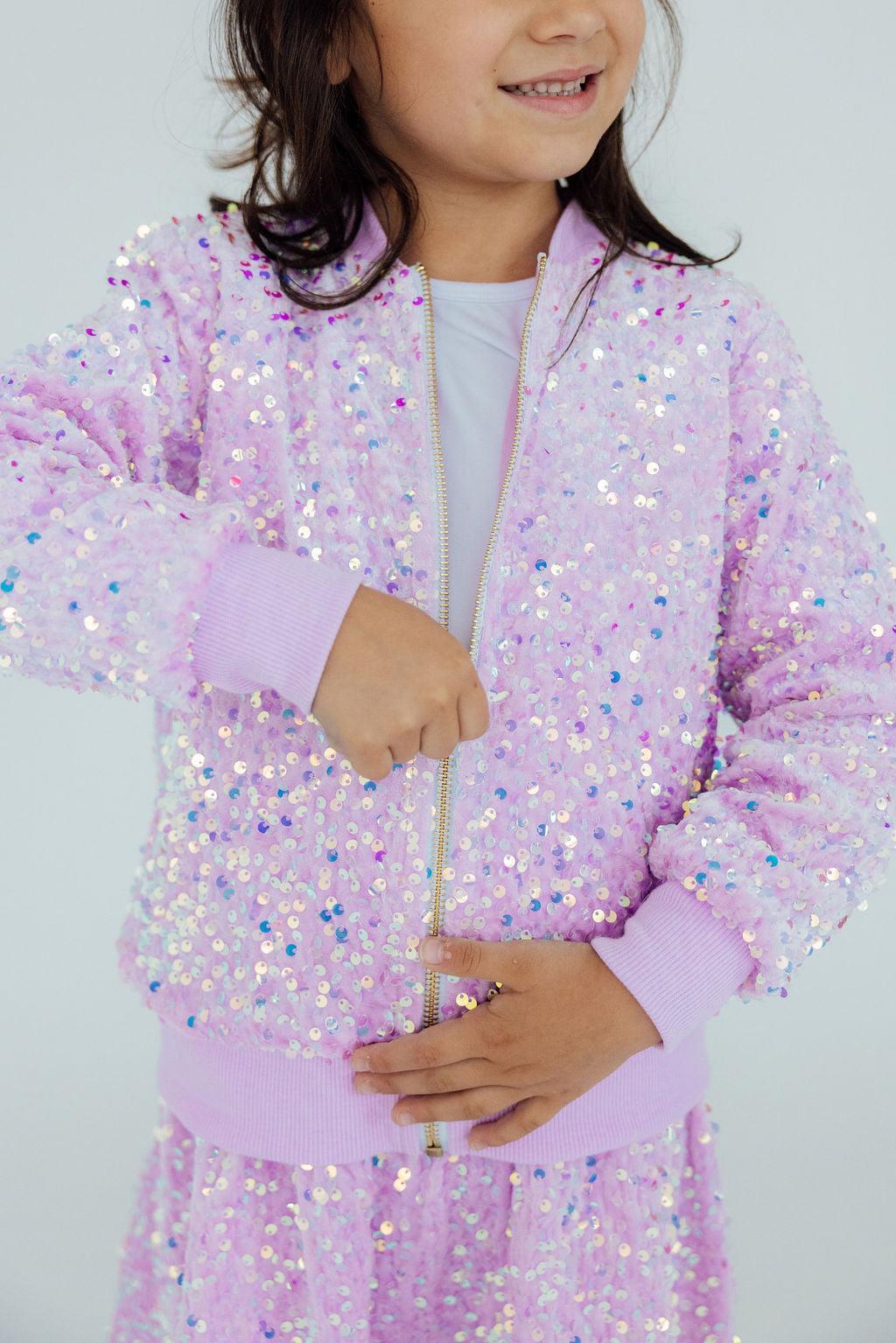 Girl wearing a Lilac Velvet Sequin Jacket from Mila & Rose, smiling and zipping up her jacket, showcasing the sparkling sequins in a stylish pose.