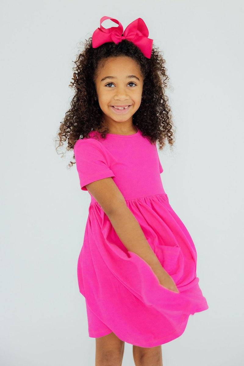 A joyful girl wearing the Hot Pink S/S Pocket Twirl Dress, smiling brightly with a big bow in her hair, ready to twirl into fun.