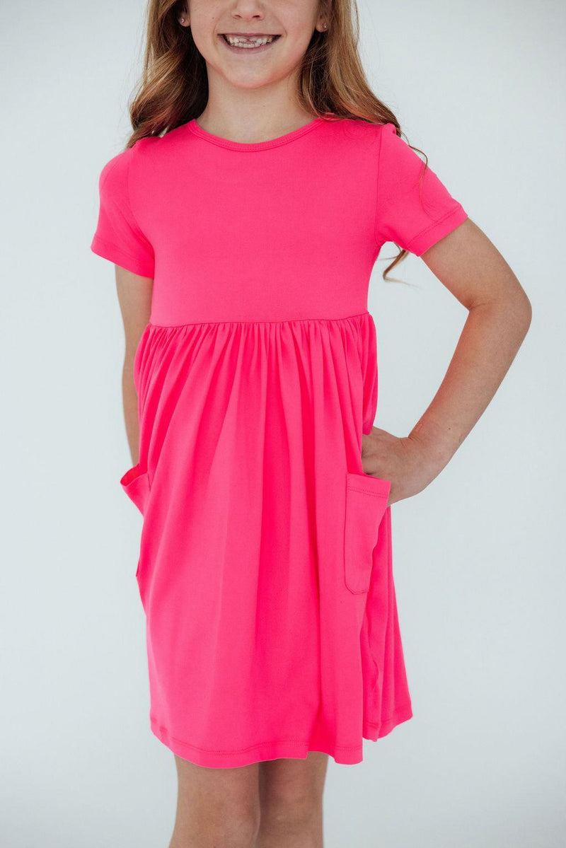 A joyful girl wearing the Neon Pink S/S Pocket Twirl Dress by Mila & Rose®, featuring a playful design with pockets, perfect for fun and active play.