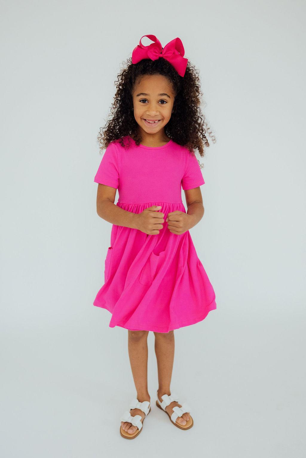 Little girl joyfully wearing the Hot Pink S/S Pocket Twirl Dress, showcasing its twirly design and vibrant color, perfect for playtime and fashion adventures!