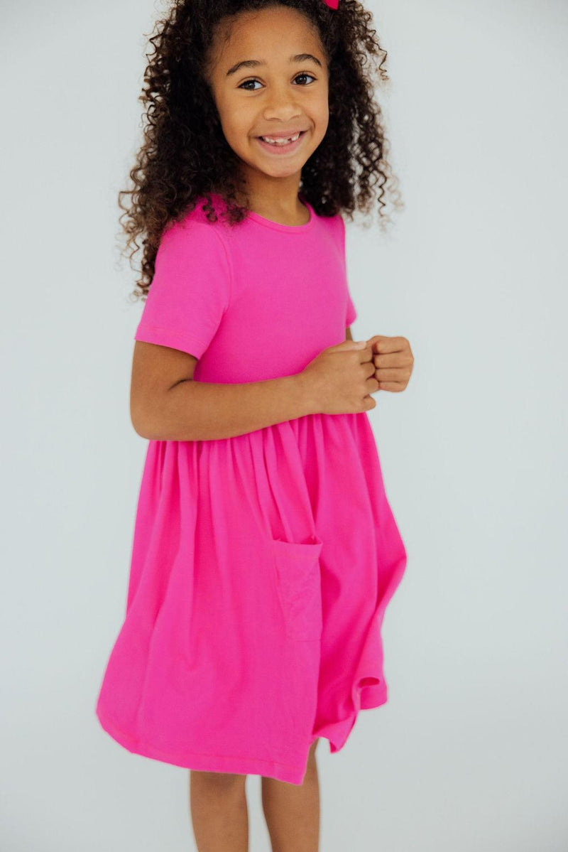 A lively girl cuddling in the Hot Pink S/S Pocket Twirl Dress, showing off her playful personality with a wide smile, perfect for all-day comfort.