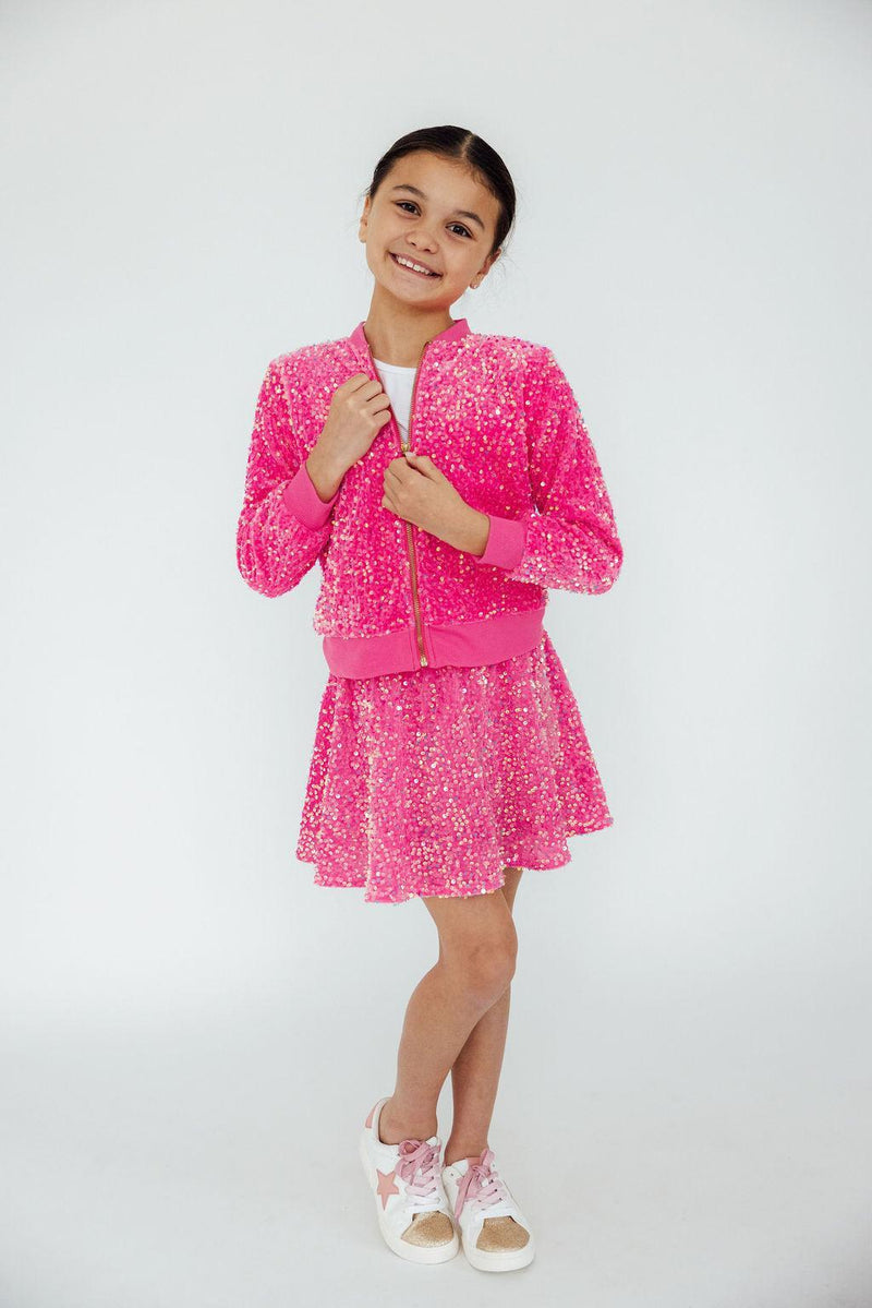 A smiling girl wearing a Pink Velvet Sequin Twirl Skort paired with a matching sequined bomber jacket, showcasing the playful design perfect for twirling and sparkling on any occasion.