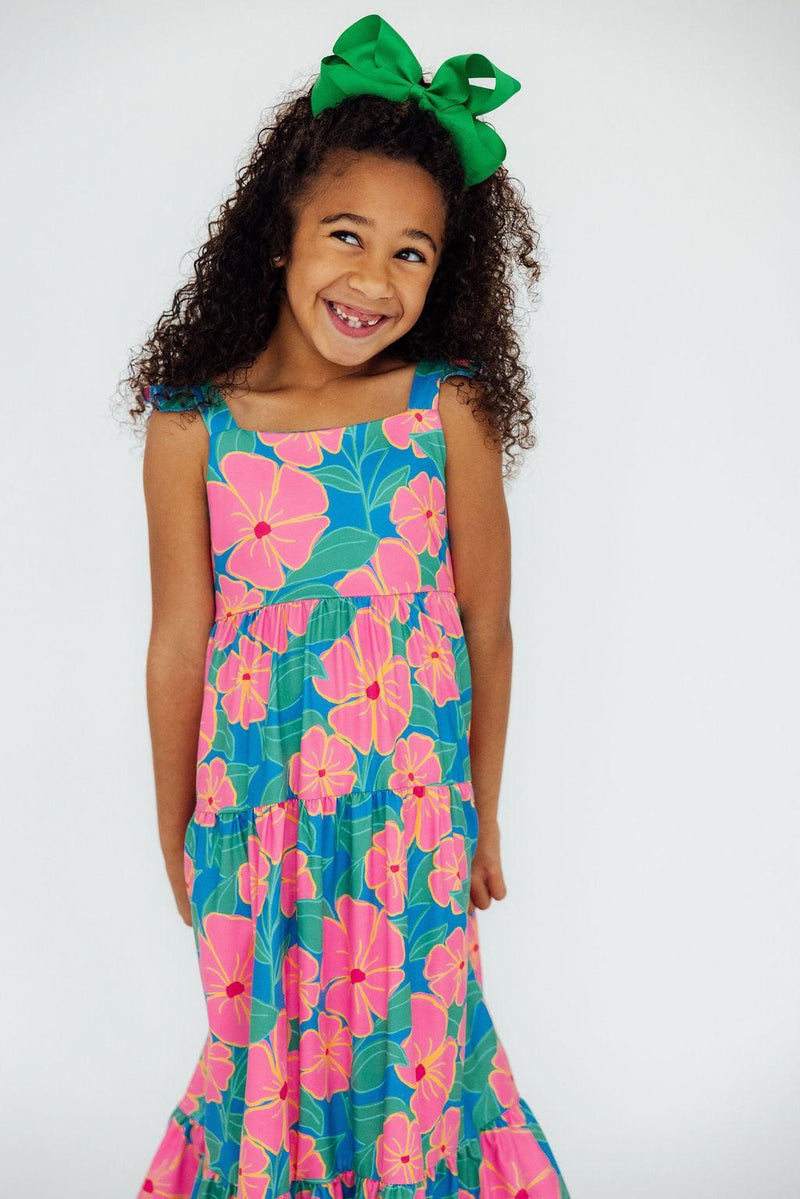 A cheerful girl wearing the Luau Floral Ruffle Maxi Dress, featuring vibrant pink and blue floral patterns, smiles at the camera with a playful green bow in her hair.