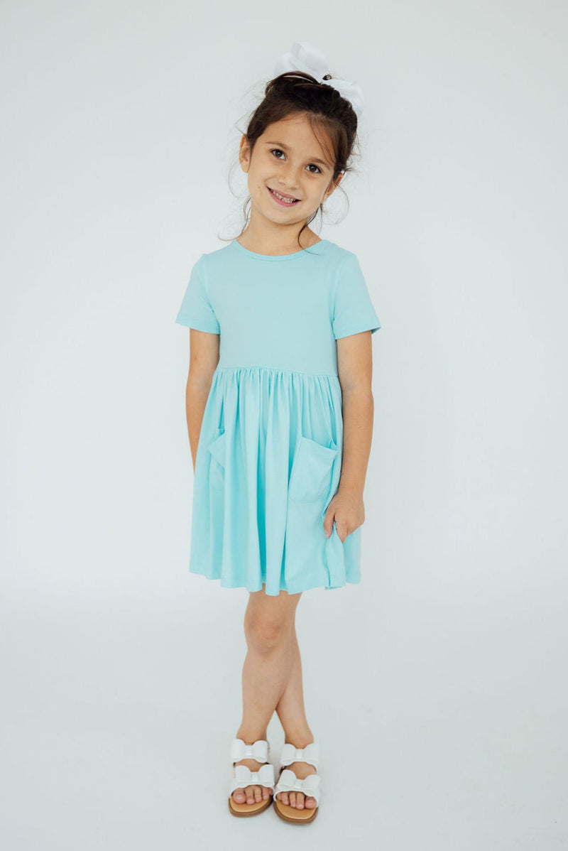 Front view of a girl wearing the Aqua S/S Pocket Twirl Dress, featuring a comfortable fit and playful twirl design, ideal for everyday adventures.