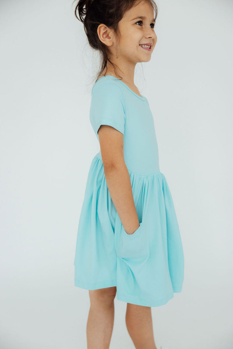 Side view of a girl happily wearing an Aqua S/S Pocket Twirl Dress, featuring a comfortable fit and playful twirl.