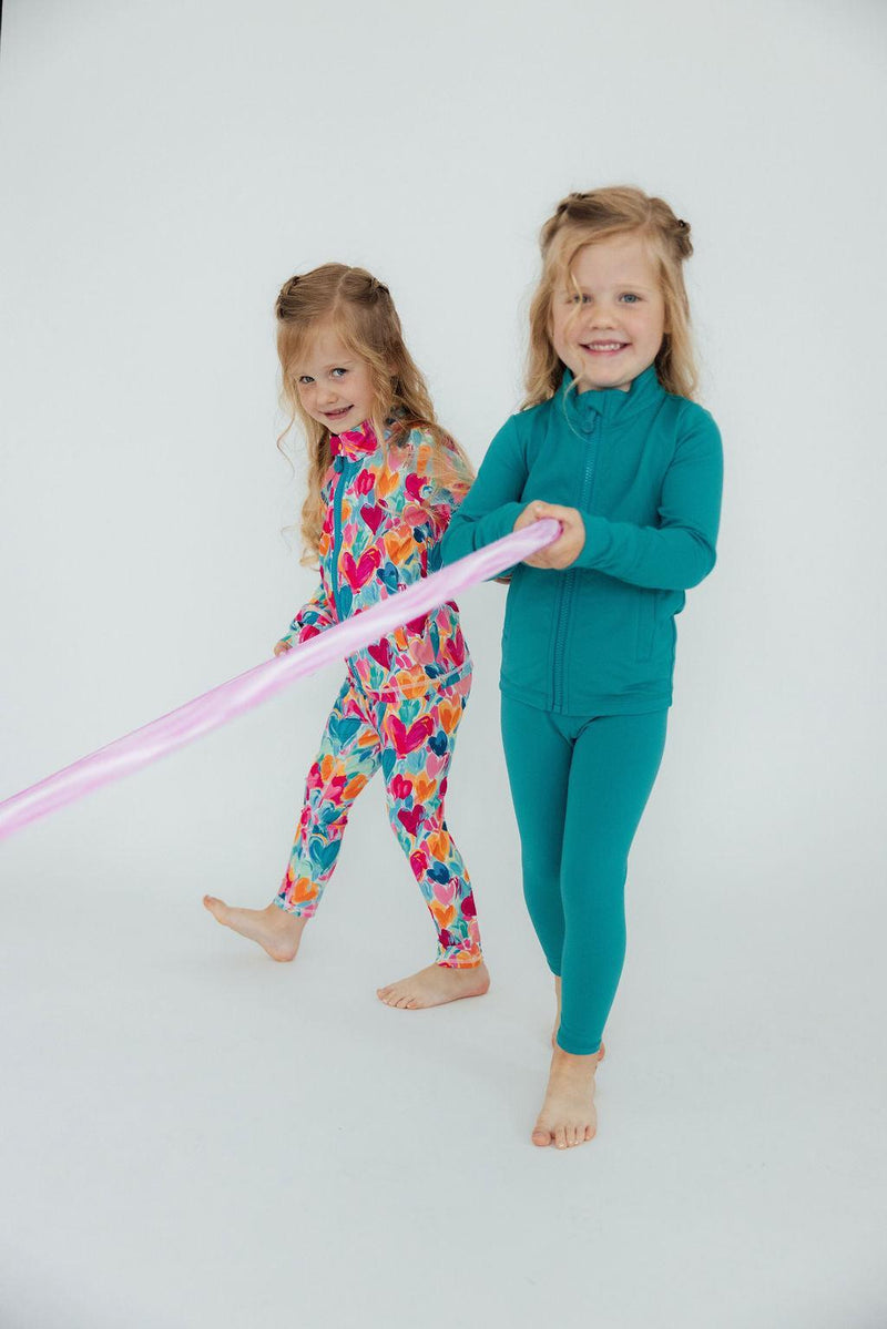 Two happy girls wearing colorful outfits, one in floral and the other in Turbo Teal Active Leggings for Girls, playing together with a ribbon, highlighting fun active fashion.