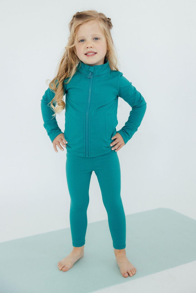 Cute girl in a teal jacket and Turbo Teal Active Leggings for Girls poses confidently on a yoga mat, representing stylish and functional active leggings for girls.