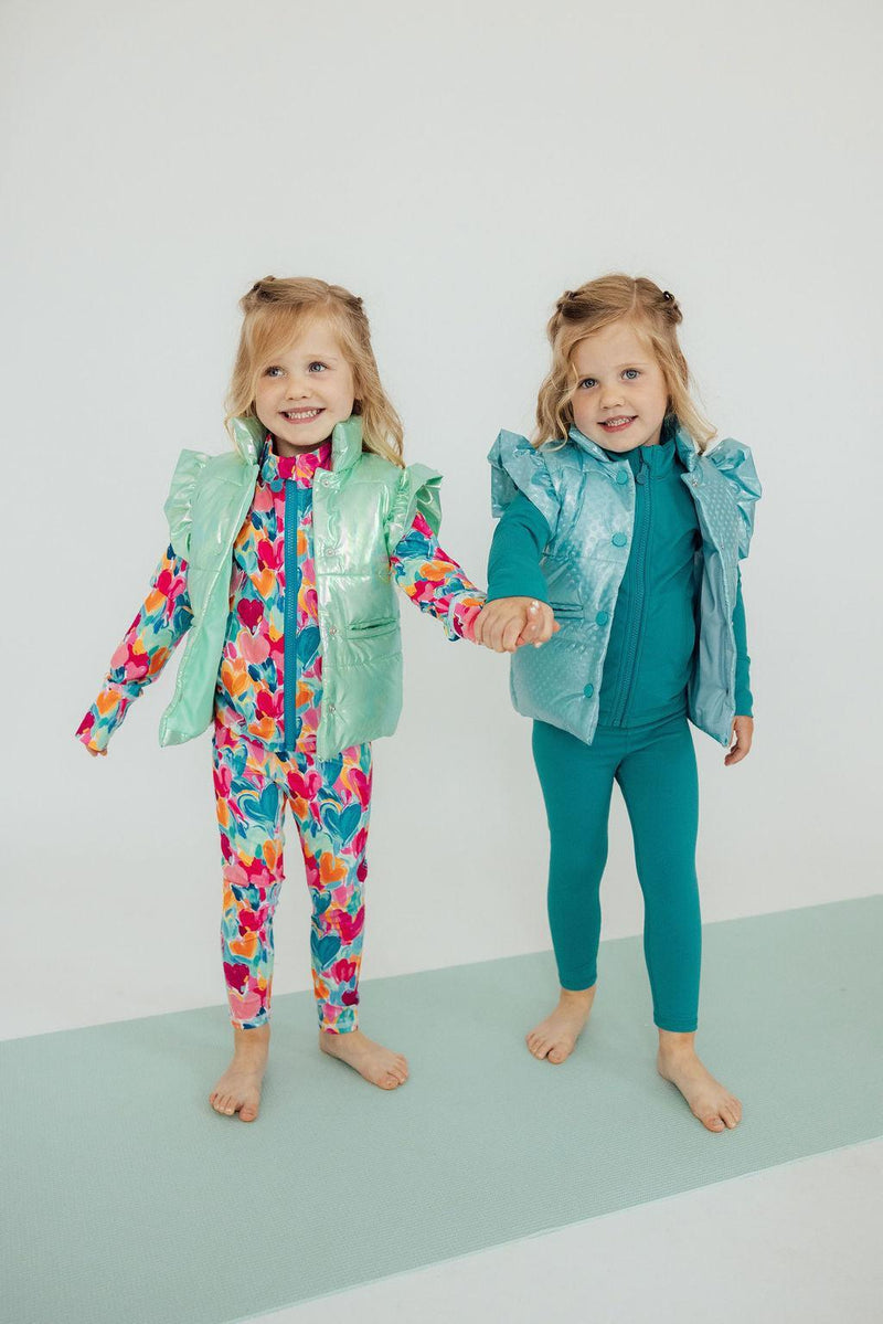 Two happy girls holding hands, one in a colorful floral outfit and the other in a Turquoise Dot Puffer Vest by Mila & Rose, highlighting fun winter fashion.