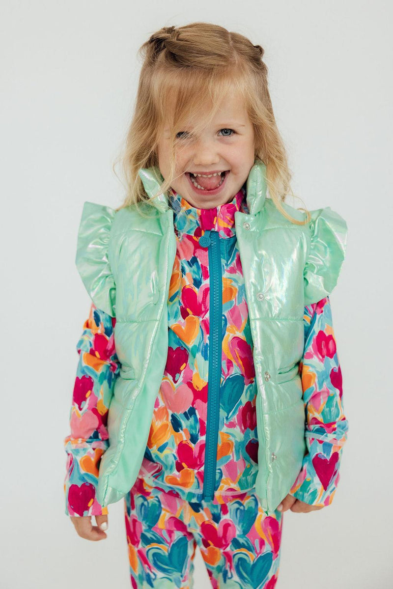 A happy girl in a Cozy Warm Mint Iridescent Puffer Vest for Girls, smiling widely while dressed in a colorful heart-patterned outfit. A great depiction of the playful mint puffer vest girls.