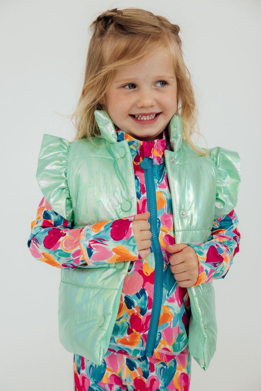 A cheerful girl wearing a Cozy Warm Mint Iridescent Puffer Vest for Girls, featuring playful ruffles and bright floral patterns underneath. Perfect for showcasing a stylish mint puffer vest girls.