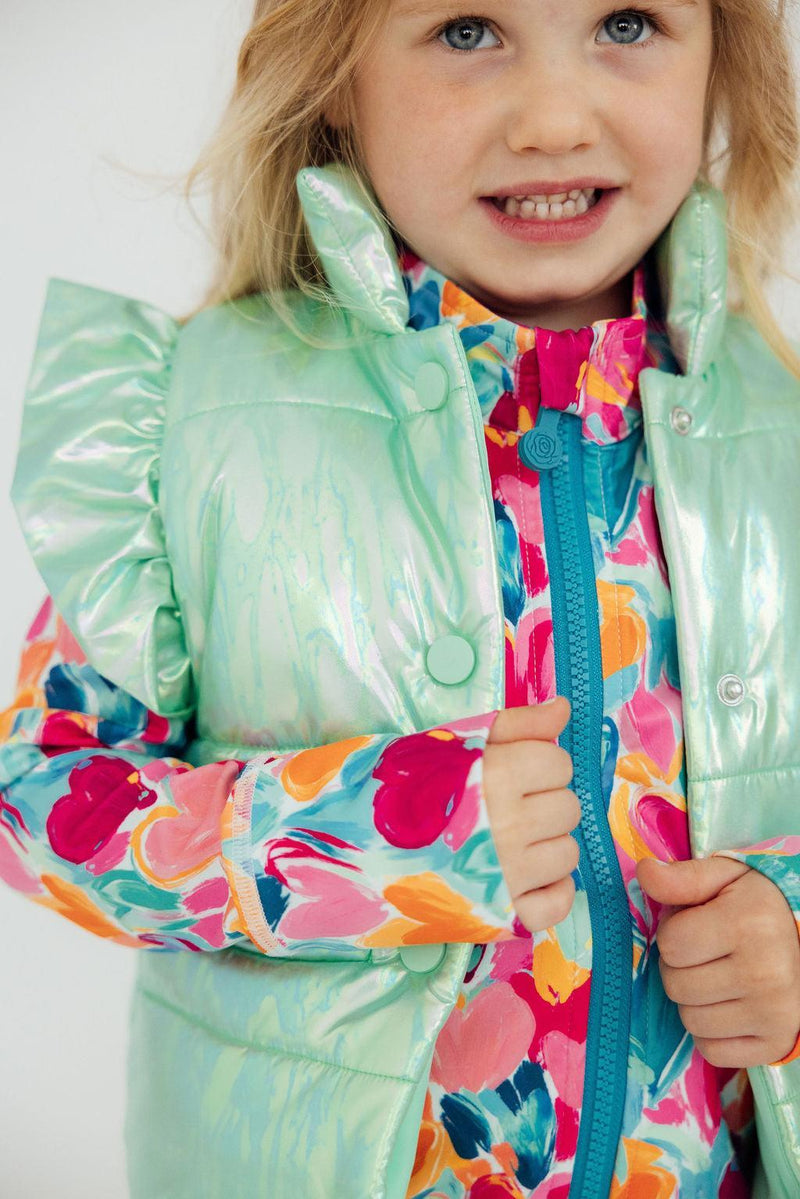 A close-up of a girl smiling while wearing a Cozy Warm Mint Iridescent Puffer Vest for Girls layered over a colorful floral outfit. Highlights the trendy mint puffer vest girls can enjoy.