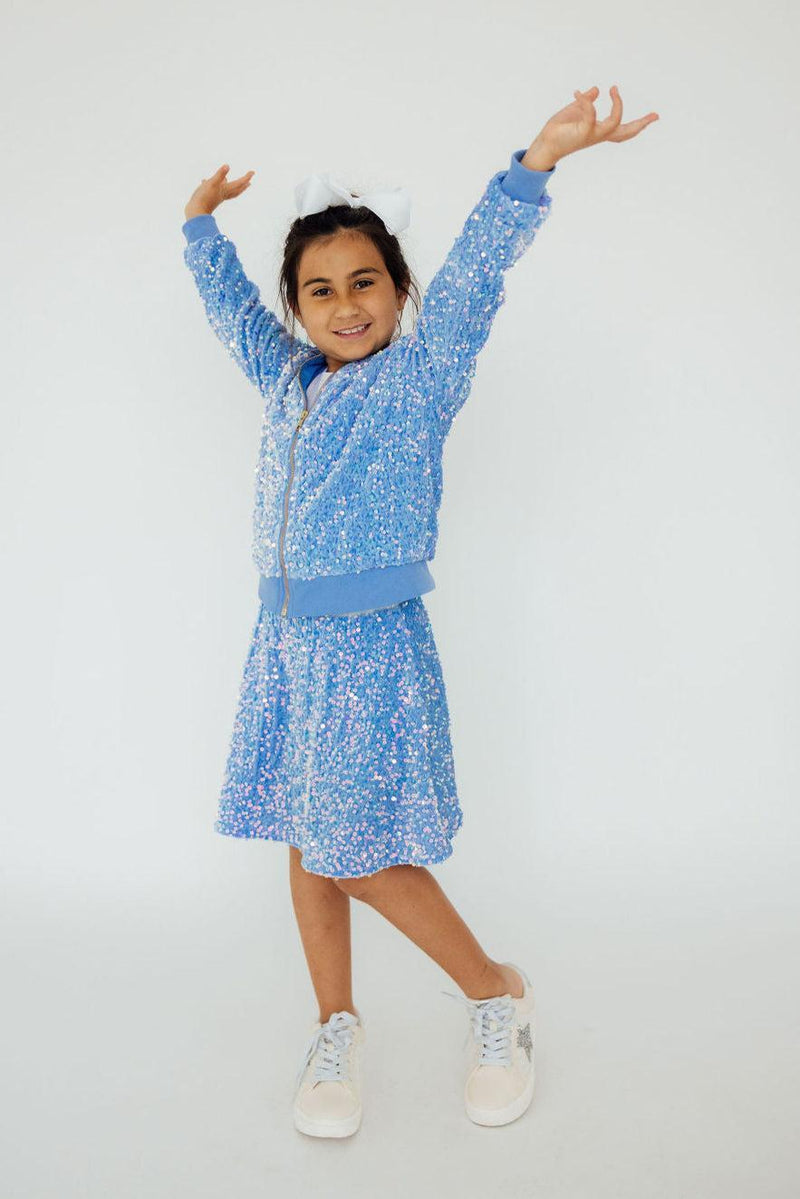 Happy girl posing with arms raised in a playful manner, wearing the Light Blue Velvet Sequin Twirl Skort from Mila & Rose and a sequined jacket.