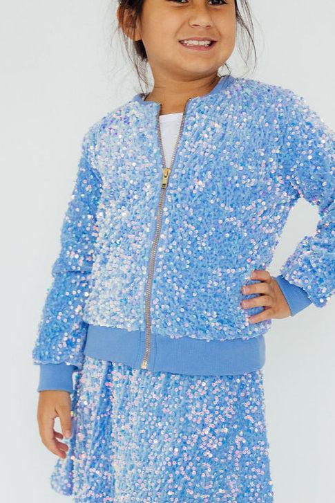 A smiling girl showcases her Light Blue Velvet Sequin Jacket by Mila & Rose ®, adorned with shimmering sequins that catch the light. The jacket's vibrant color adds a playful touch, making it an ideal choice for both festive occasions and casual outings.