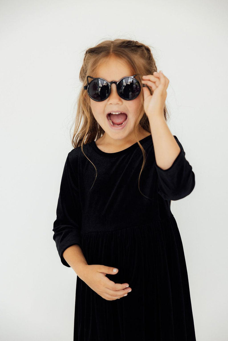 Happy girl in the SALE Black Velvet Twirl Dress by Mila & Rose®, playfully adjusting stylish sunglasses, adding a fun touch to her look.