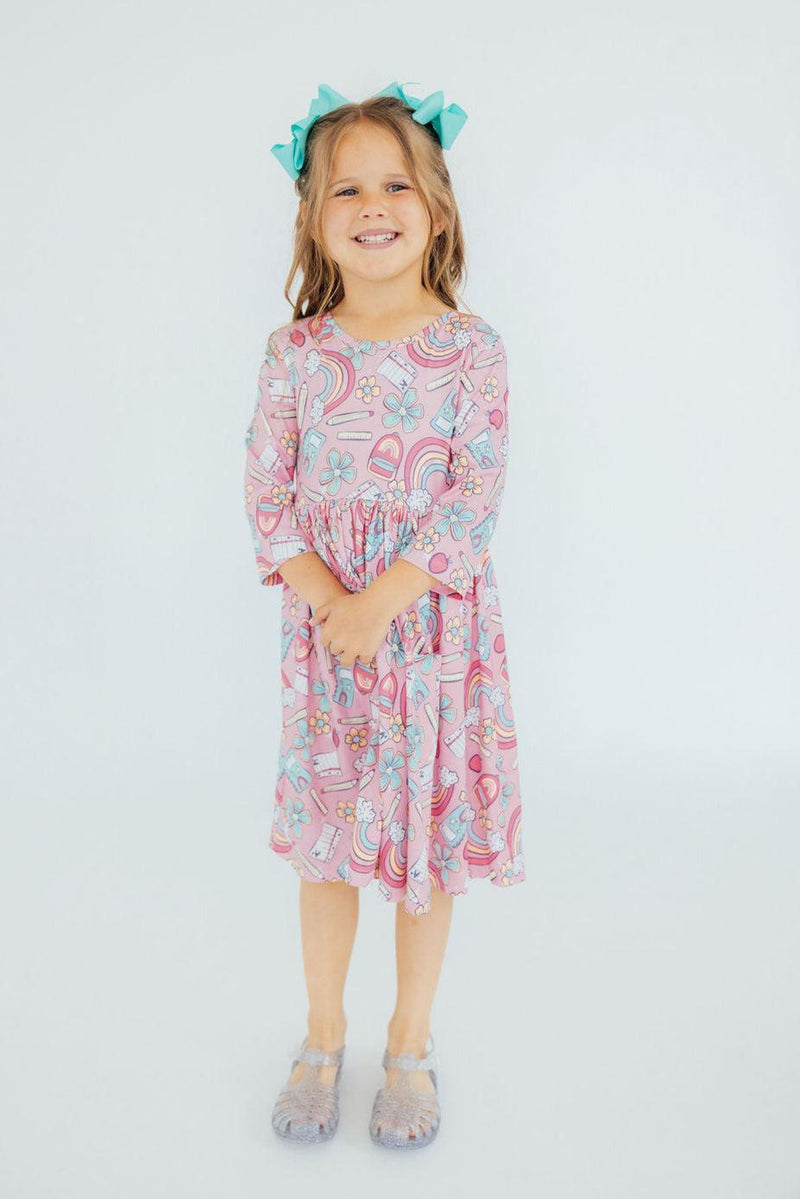 A young girl wearing the Dream Big Little One 3/4 Pocket Twirl Dress, smiling and standing happily. The dress features colorful patterns with rainbows and flowers.