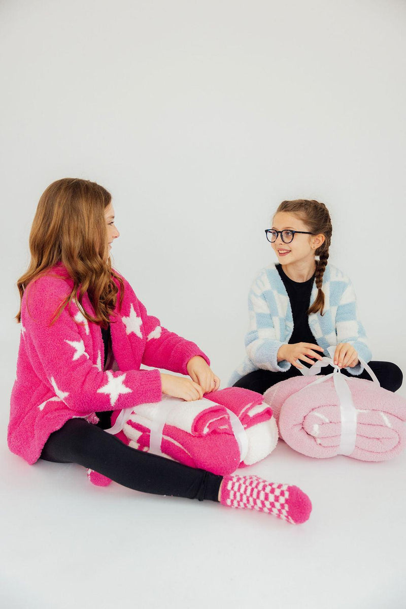 Two friends lounging with the Light Pink Cowgirl Cozy Blanket, surrounded by other cozy blankets, radiating comfort and fun.