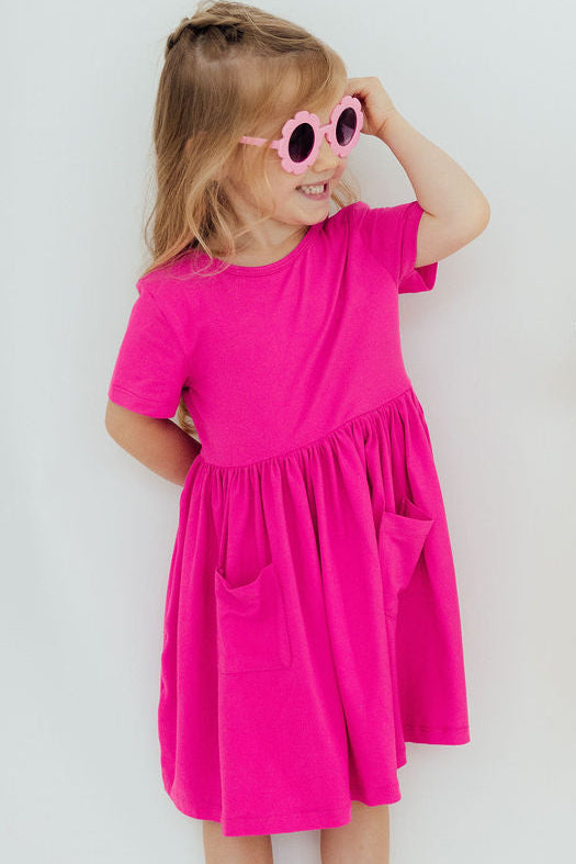 A girl in a vibrant Hot Pink S/S Pocket Twirl Dress striking a cheerful pose, wearing stylish sunglasses, perfect for sunny adventures.
