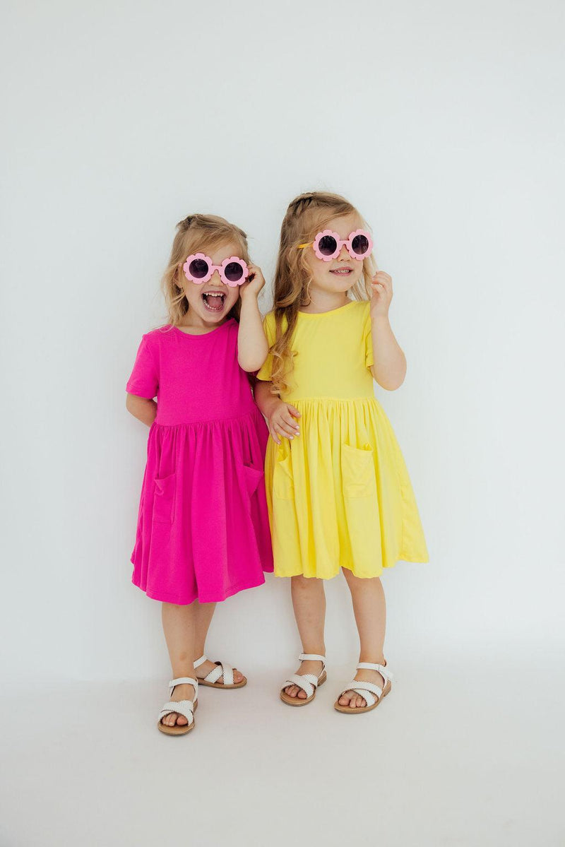 Two girls wearing the Hot Pink S/S Pocket Twirl Dress and a yellow dress, sporting playful sunglasses and enjoying a fun day of twirling.