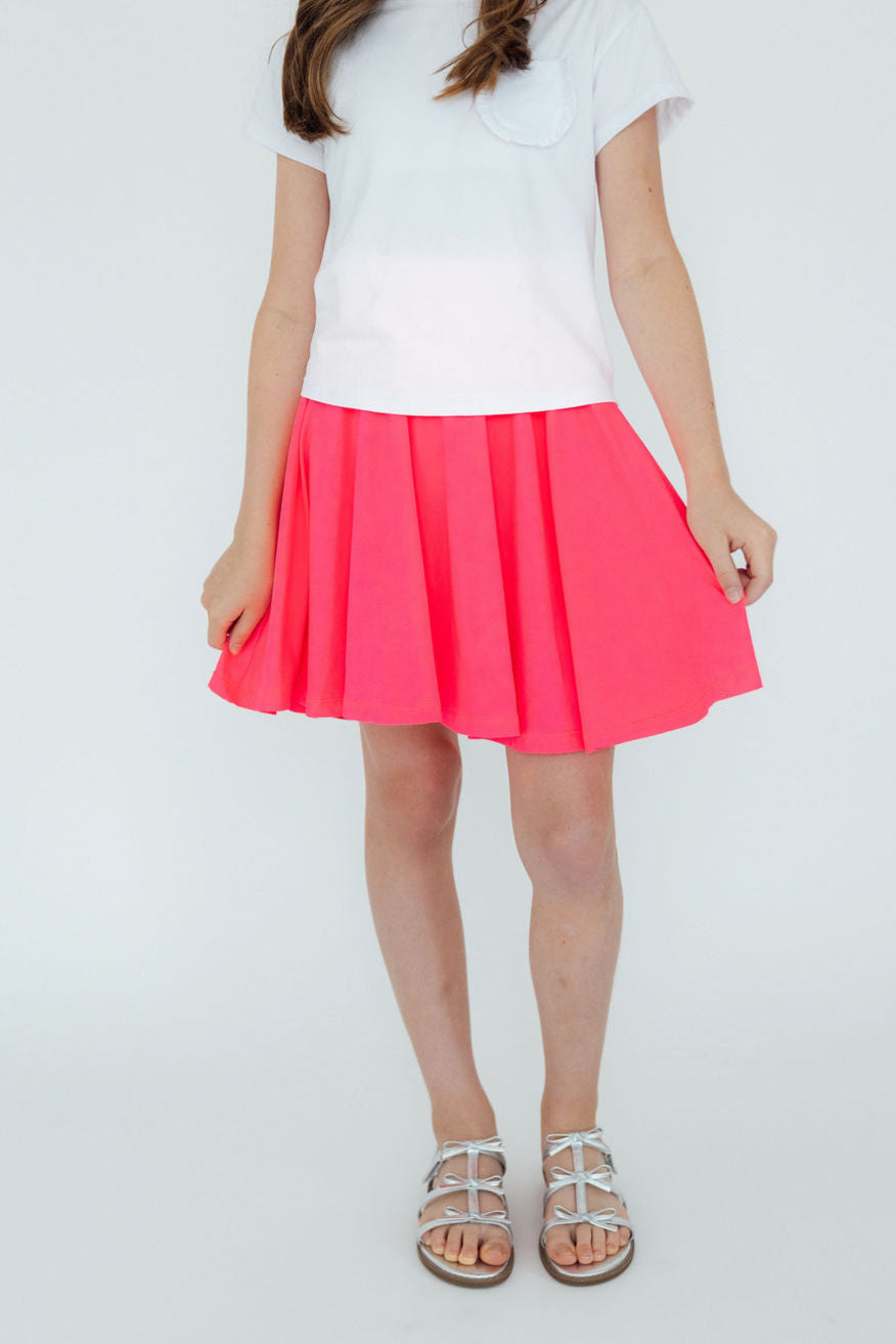 A young girl wearing a white shirt and the vibrant Neon Pink Twirl Skort, showcasing its playful pleated design and attached shorts underneath, perfect for active play.