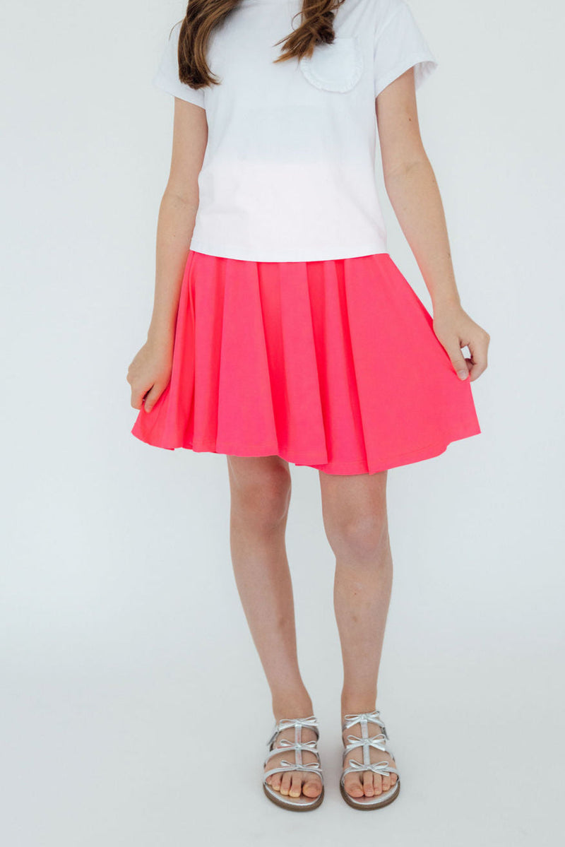A young girl wearing a white shirt and the vibrant Neon Pink Twirl Skort, showcasing its playful pleated design and attached shorts underneath, perfect for active play.