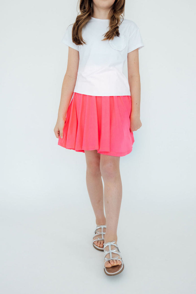 A girl wearing a Neon Pink Twirl Skort from Mila & Rose, paired with a white t-shirt, standing confidently with feet together on a light background, highlighting the playful design of the skort.