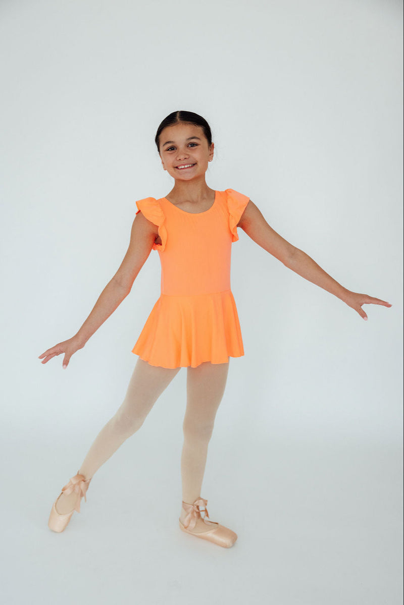 A young ballerina wearing a Neon Coral Short Sleeve Twirl Leotard by Mila & Rose, showcasing her dance pose with a joyful smile. The bright coral leotard features short flutter sleeves, ideal for dance classes or playdates, paired with classic ballet shoes.