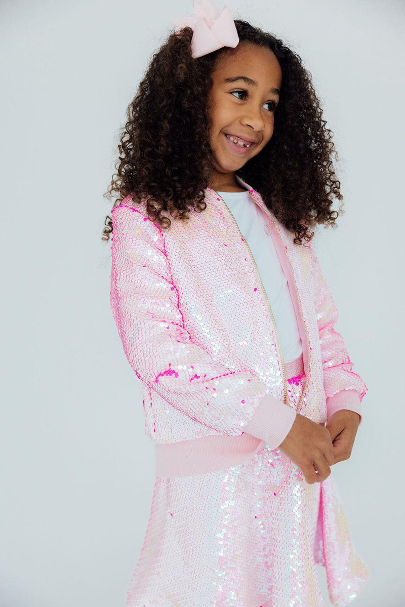 A stylish young girl wearing the Diva Pink Flip Sequin Jacket, sparkling in sequins while confidently posing, capturing the essence of fun and creativity.