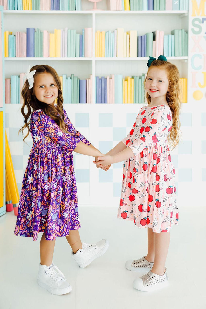 Two girls holding hands, one wearing the Star Student 3/4 Sleeve Pocket Twirl Dress in pink with apples and the other in a vibrant floral design, perfect for friends' playdates.