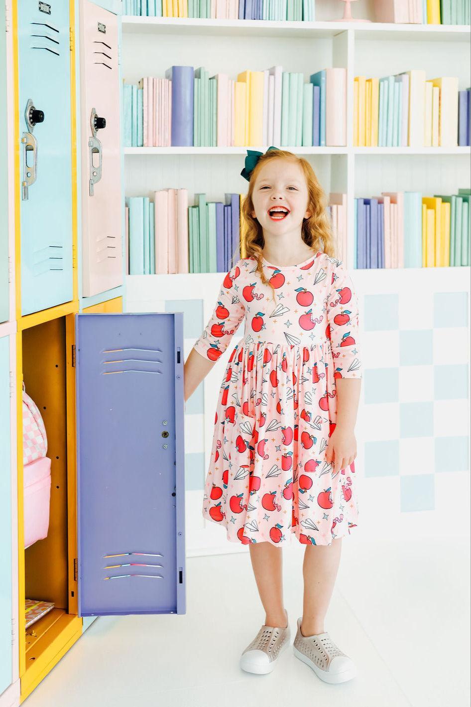 Girl smiling in a Star Student 3/4 Sleeve Pocket Twirl Dress by Mila & Rose, standing next to a pastel-colored locker, perfect for school and play.