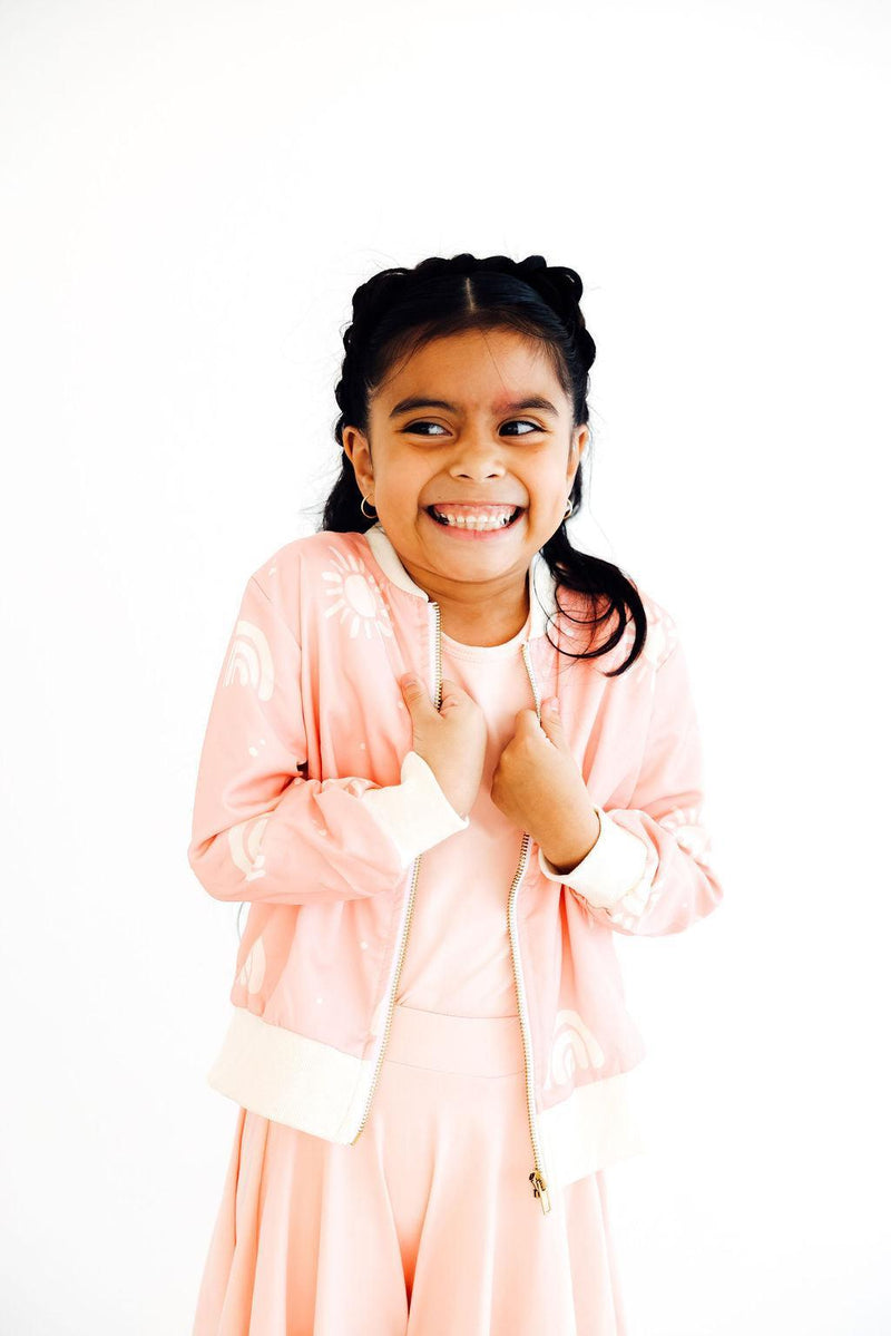 A cheerful girl with braids in the Soft Pink Sun Satin Jacket by Mila & Rose ®, holding the jacket close. She radiates joy, highlighting the jacket's fun and fashionable style.