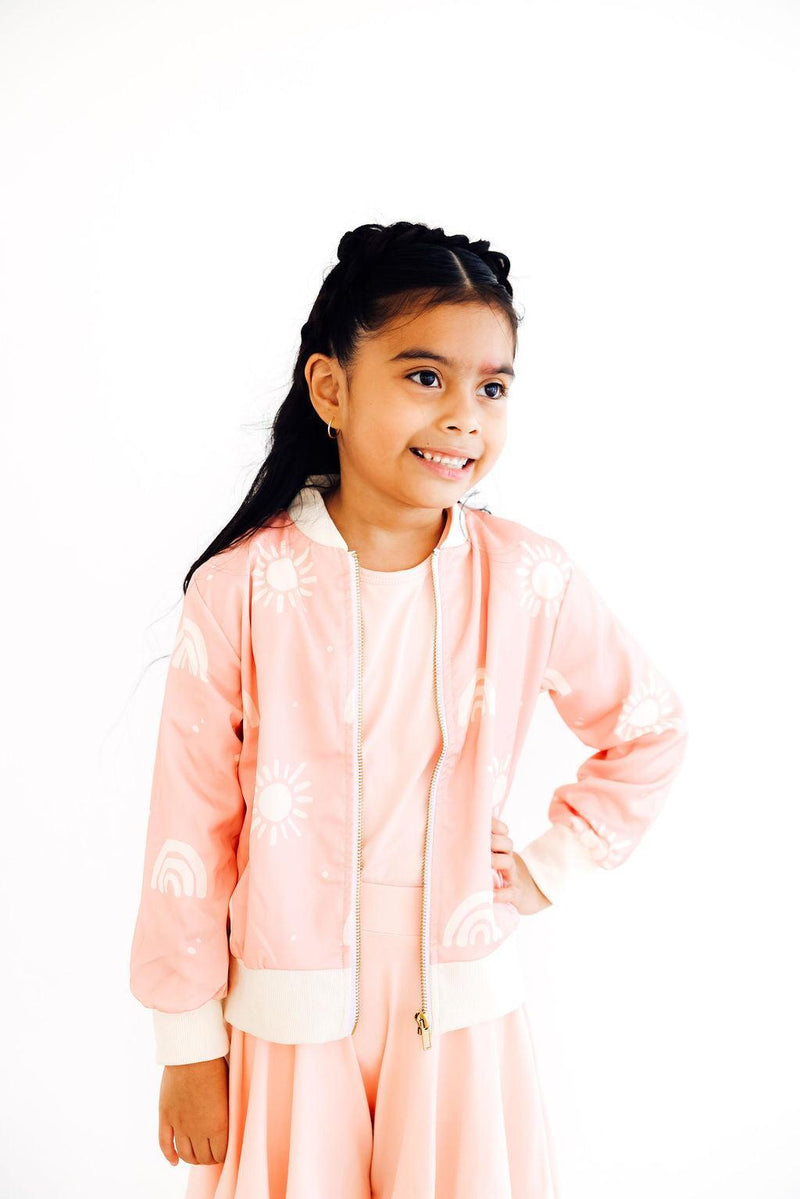 A happy young girl wearing the Soft Pink Sun Satin Jacket by Mila & Rose, smiling while looking to the side. The charming jacket adds a touch of elegance to her playful outfit.